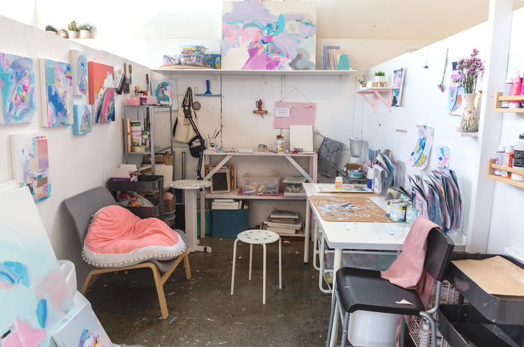 An artist studio at Akin Dupont with many soft pastel coloured abstract paintings on the walls, tables covered in art supplies, two chairs and a stool. 