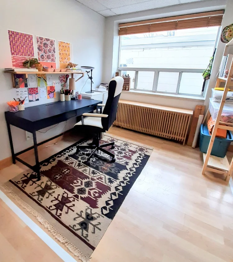  A light-filled Davisville studio features a large window at the back with a sizable radiator beneath it. Her desk and chair, along with textile patterns affixed to the wall, are positioned along the left-hand side, while a tall standing shelf occupi
