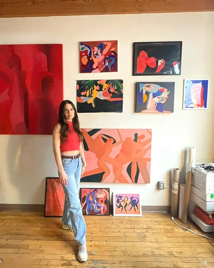 Artist and Akin Member Franziska standing in her Akin Richmond Bathurst studio weaning a red top and light blue jeans and looking forward with a slight smile. Behind her are a series of paintings in various sizes.

