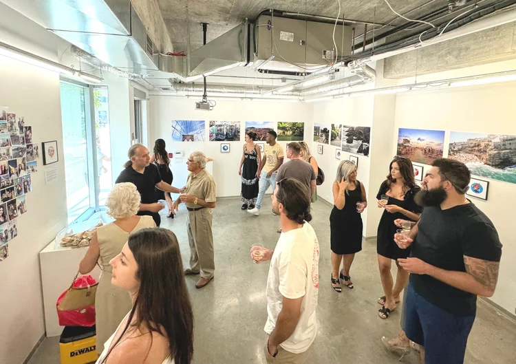 A small group of people in Remote gallery, some are talking together some are looking at aart on the walls, they are in various areas of the gallery. The gallery has white walls, and colourful artwork posted on the walls. 