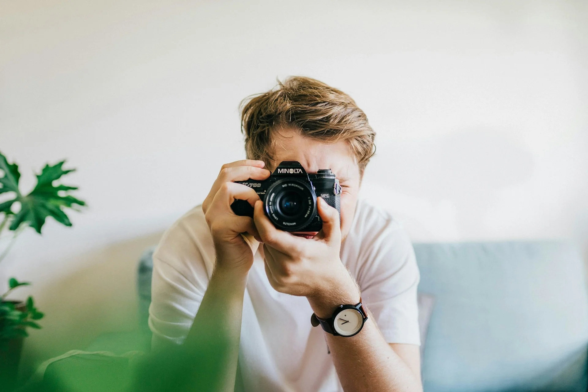 A person holding a camera up to their face, taking a photo indoors with plants in the background.