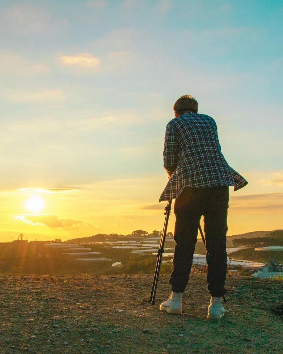 Person standing on a hill at sunset, looking out over a landscape while holding a tripod.