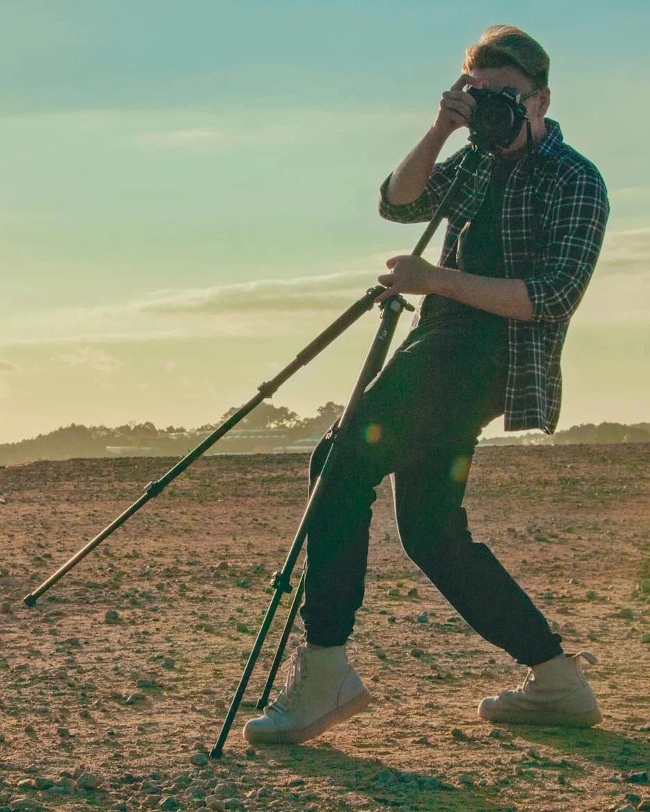 Photographer leaning into a tripod-mounted camera on a rocky landscape, silhouetted by warm sunset light.