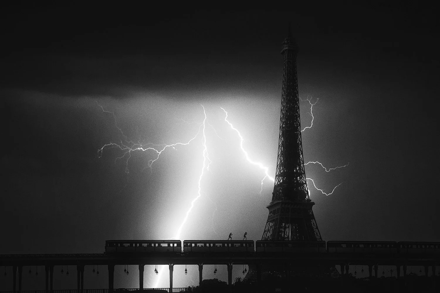 Black and white photograph of lightning striking beside the Eiffel Tower as a train crosses a bridge