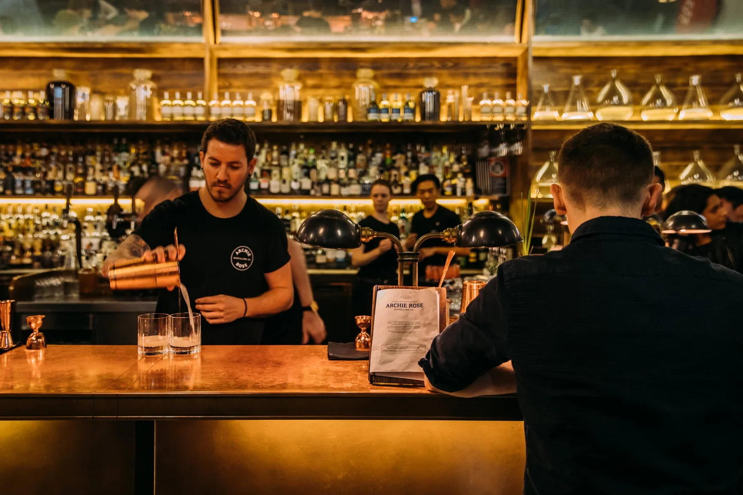 A bartender pouring a spirit at the Archie Rose bar in Rosebery, the back bar lined with bottles and a guest waiting at the counter
