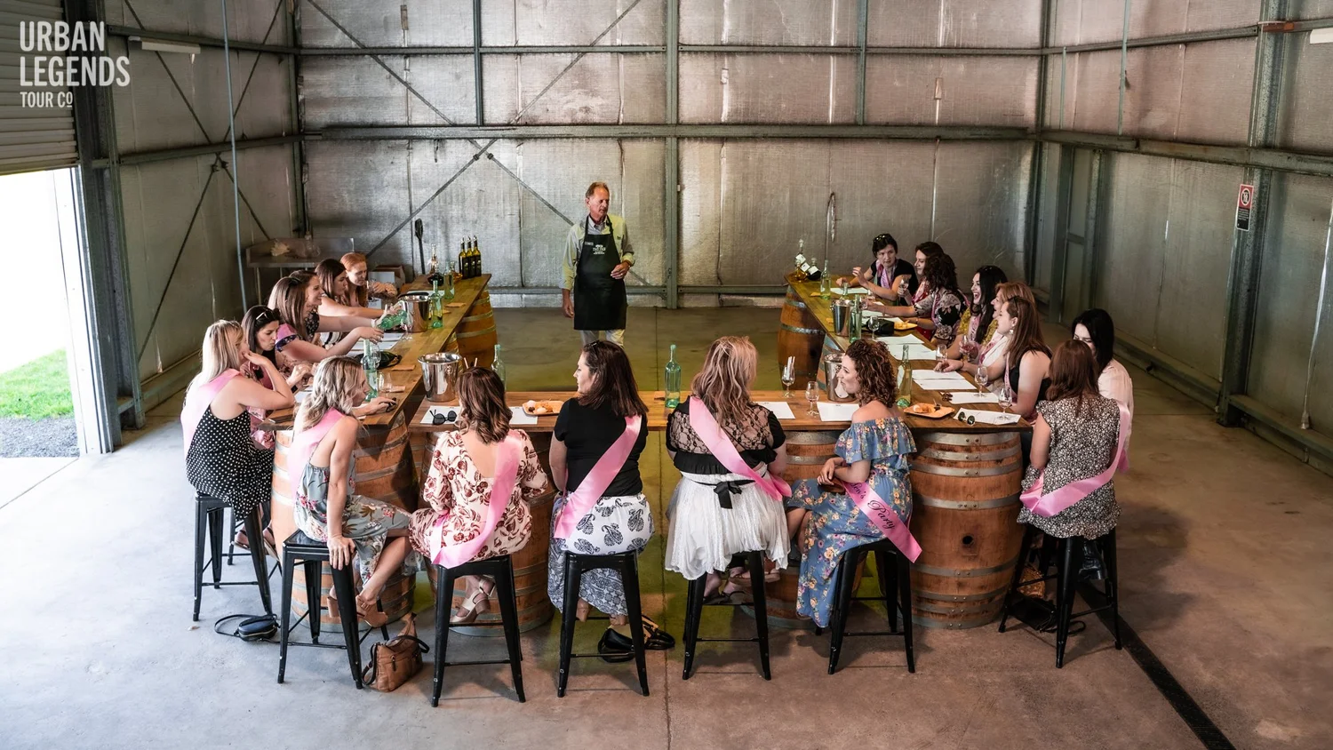 A hens party of around sixteen guests in pink sashes seated at barrel tables while a winemaker presents a tasting in a Hunter Valley cellar door barn.