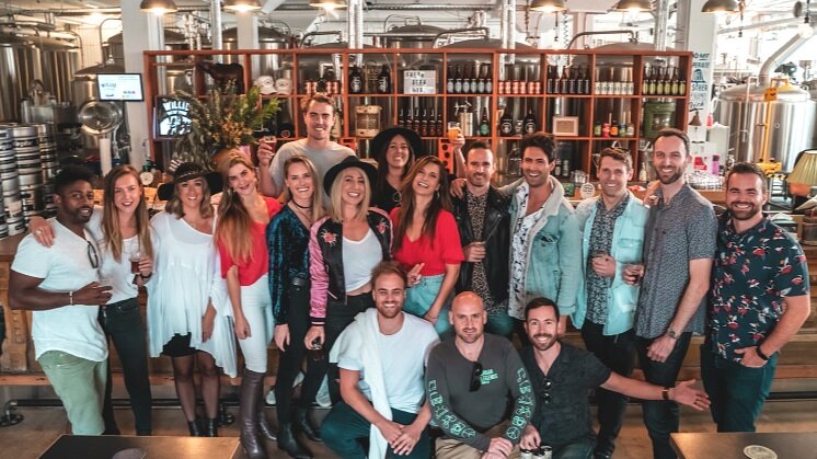 An Urban Legends group of around eighteen posed together inside a Marrickville brewery, fermentation tanks and a bottle shelf behind them
