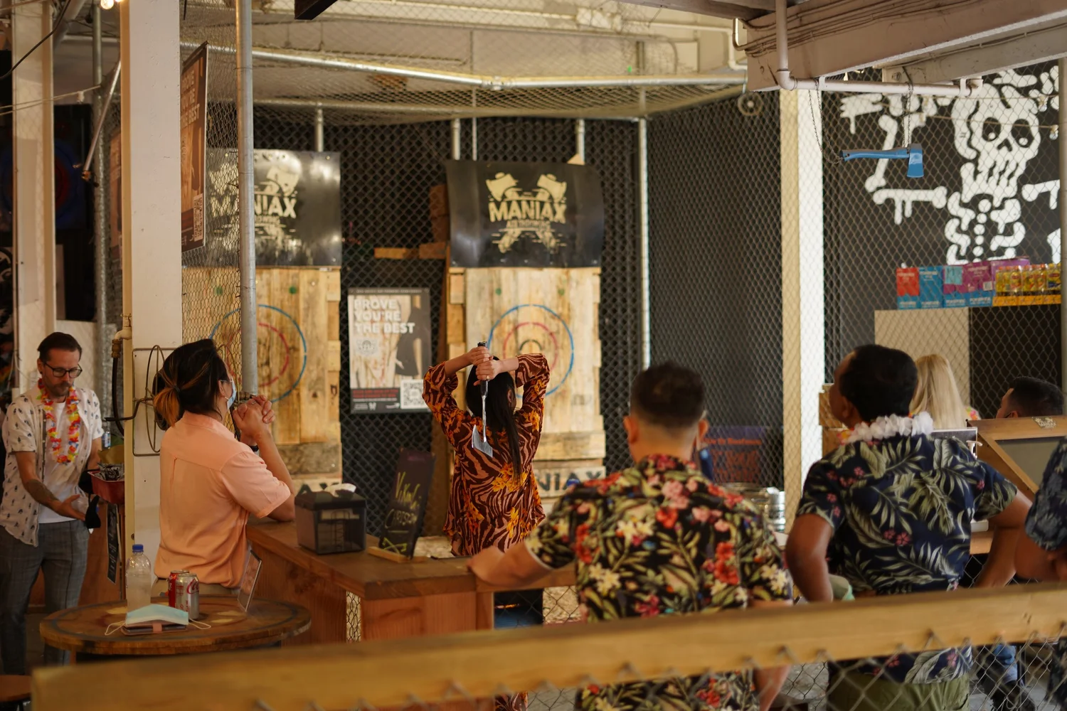 A group in bright shirts throwing axes at timber targets inside a Maniax axe throwing venue, caged lanes and scoreboards along the wall