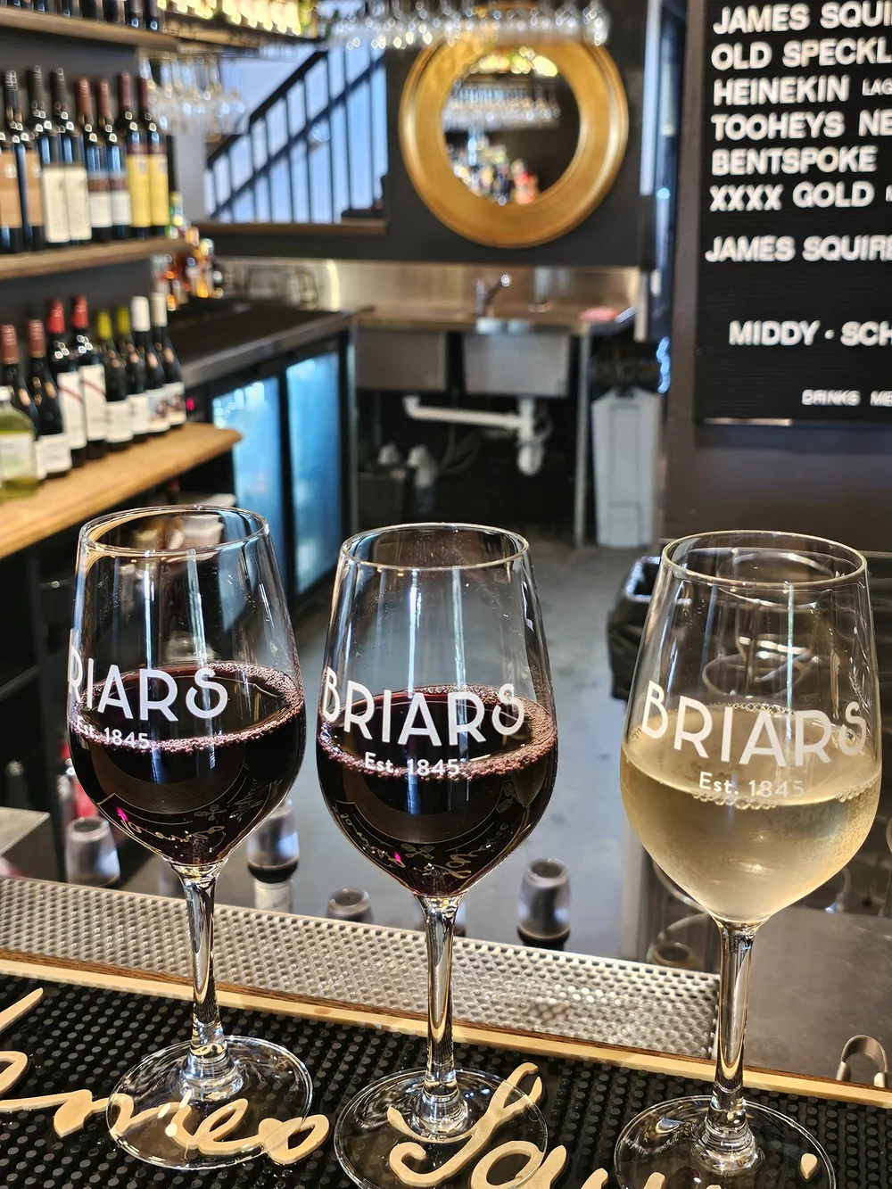 Three Briars Est. 1845 tasting glasses on a bar, two of red wine and one of white.
