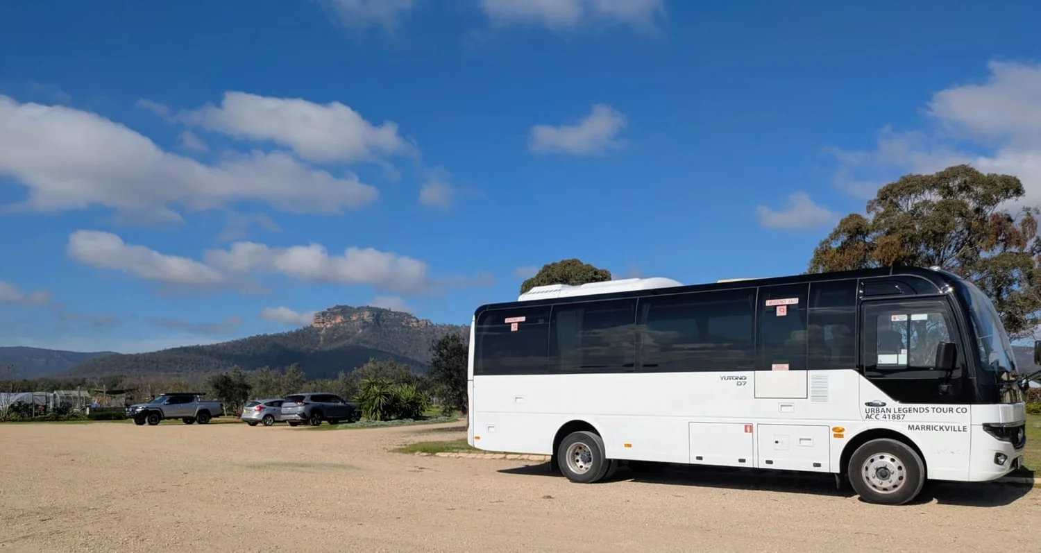 A white Urban Legends 27 seat coach in side profile at a Hunter Valley cellar door with the escarpment behind, on a half day wine and brewery tour.