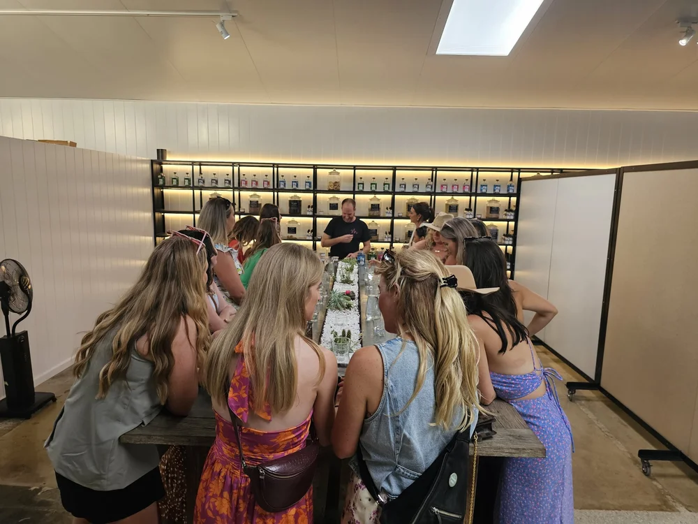 A group gathered along a long tasting table in front of a backlit shelf of gin bottles and botanical jars, a host pouring at the far end.