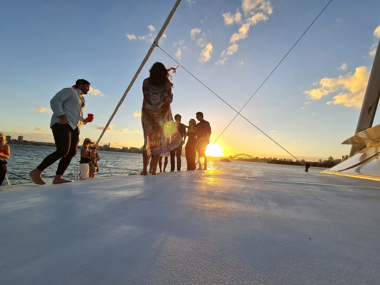 Guests standing in silhouette along the wide white foredeck of a charter yacht as the sun sets over Sydney Harbour, with the Harbour Bridge low on the horizon