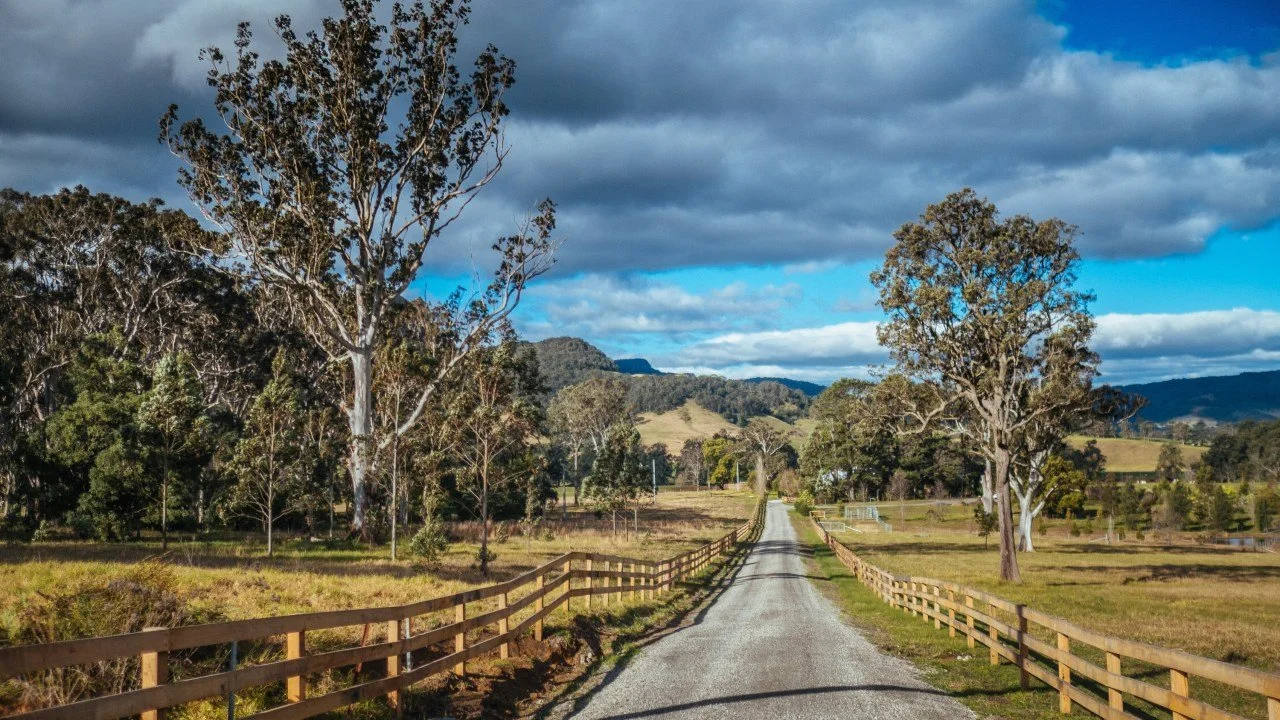 A gravel country road running between timber post-and-rail fences towards green Southern Tablelands hills under heavy cloud