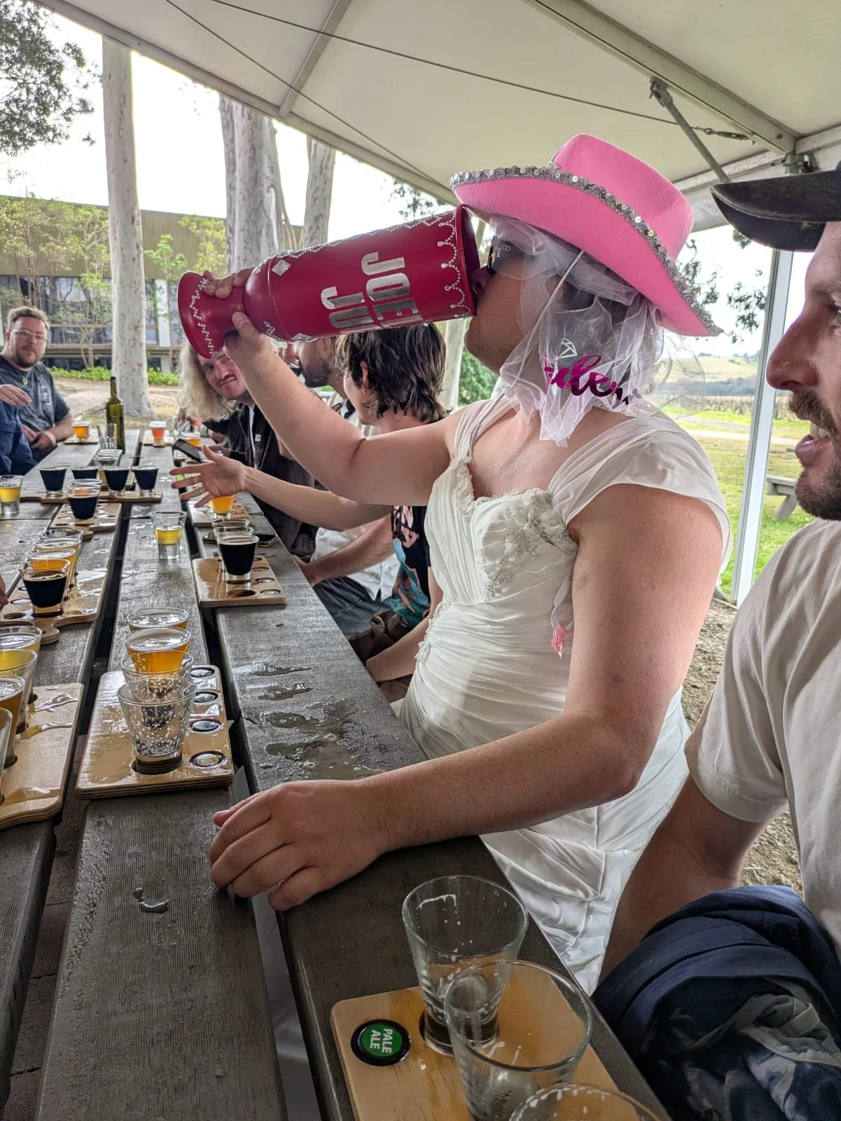 Bucks party group seated at a long table under a marquee with beer tasting paddles, vines and gum trees behind, Hunter Valley
