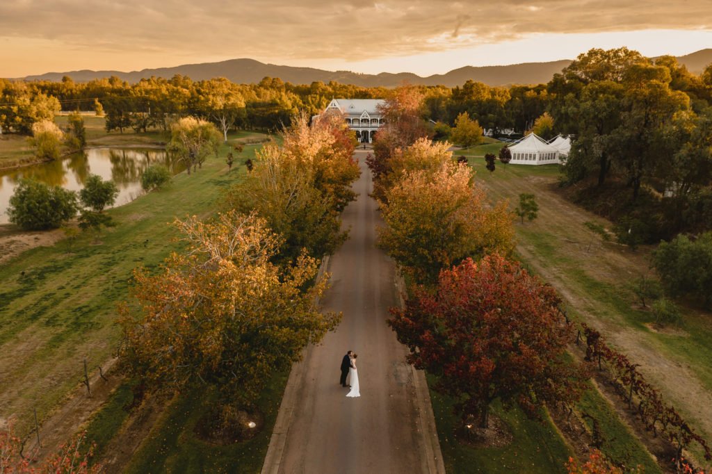 Aerial view of a bride and groom standing together on a long tree-lined driveway at a Hunter Valley estate, autumn colour on both sides and a marquee beyond the trees