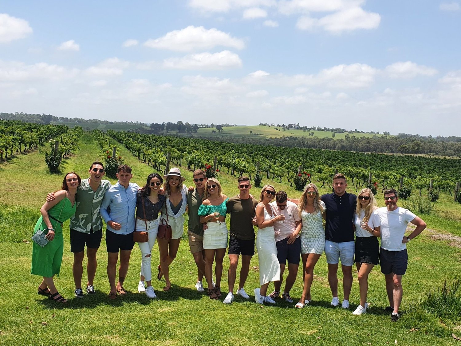 Twelve guests standing together in the vine rows on a clear day in the Hunter Valley, partway through a combined winery, brewery and distillery tour.