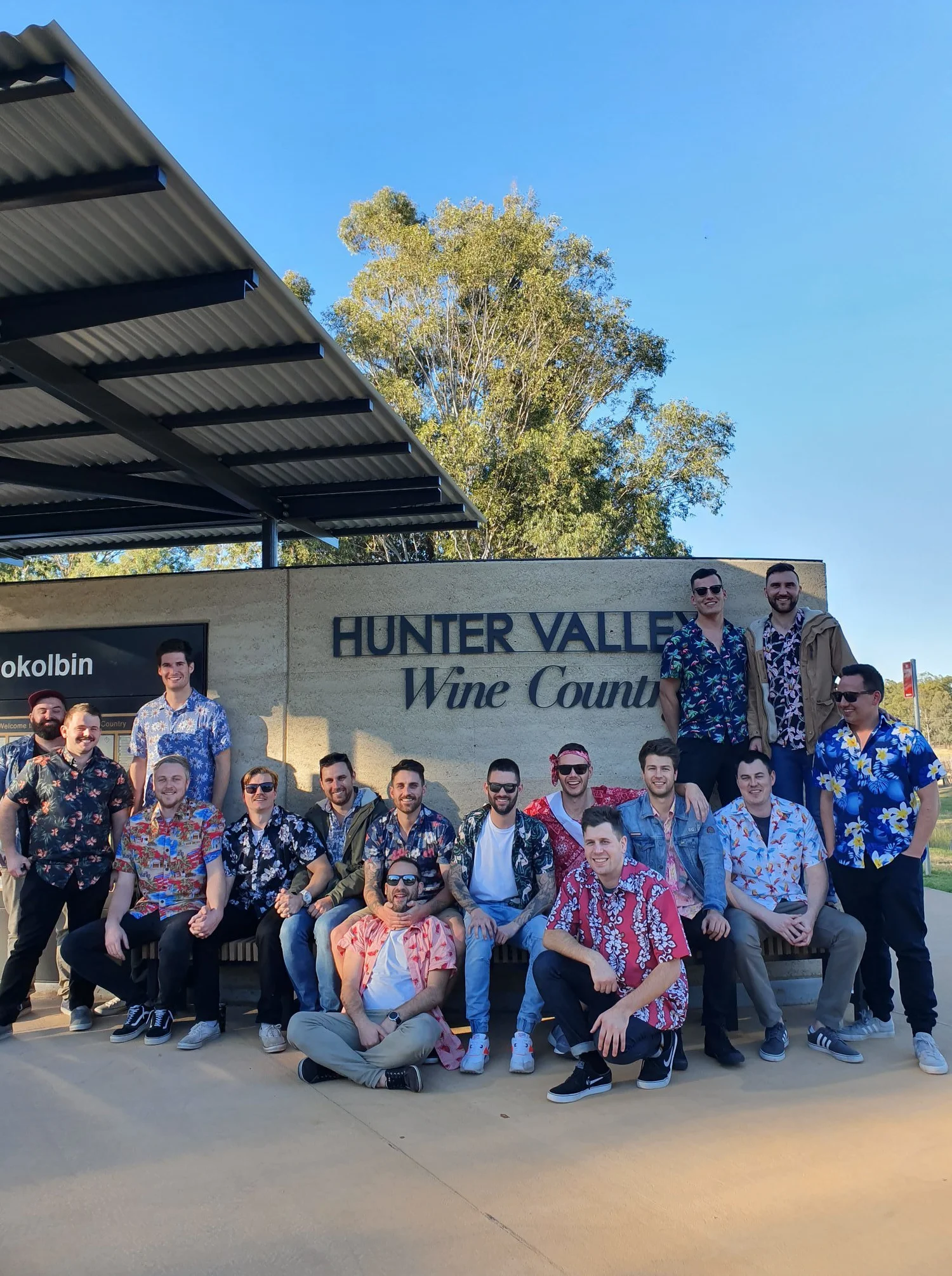 A group of fifteen guests in matching shirts gathered under the Hunter Valley Wine Country sign at Pokolbin before boarding for a brewery tour.