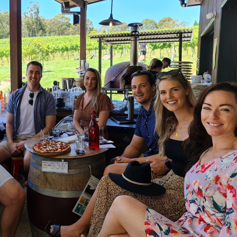 Winery, brewery and distillery tour group at a Southern Highlands cellar door Five guests seated around a wine-barrel table with a wood-fired pizza and a bottle of rosé, vineyard rows behind them.