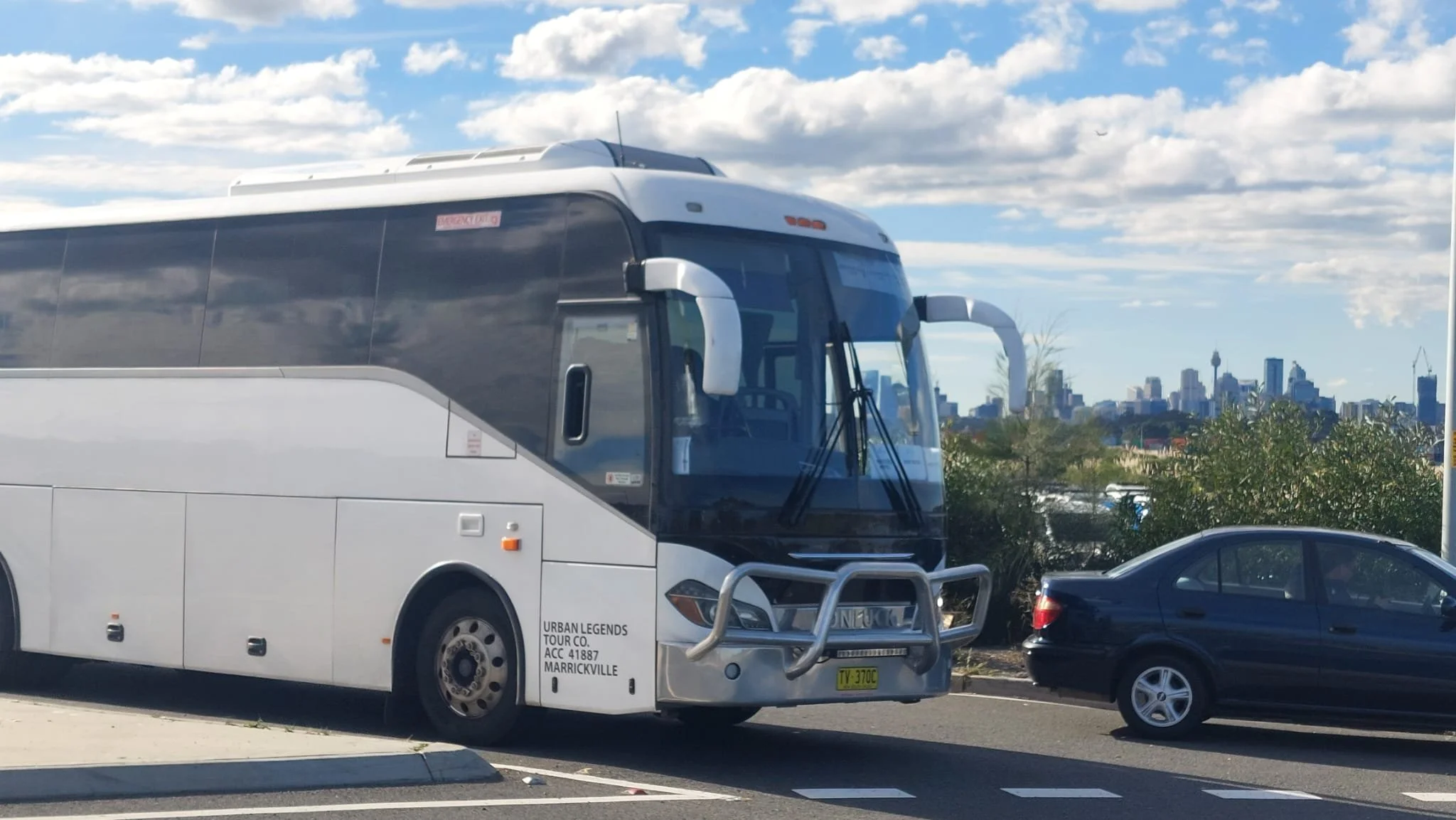 A white Urban Legends 41 seat luxury coach with silver bull bar, Urban Legends Tour Co and accreditation 41887 on the panel, Sydney skyline behind.
