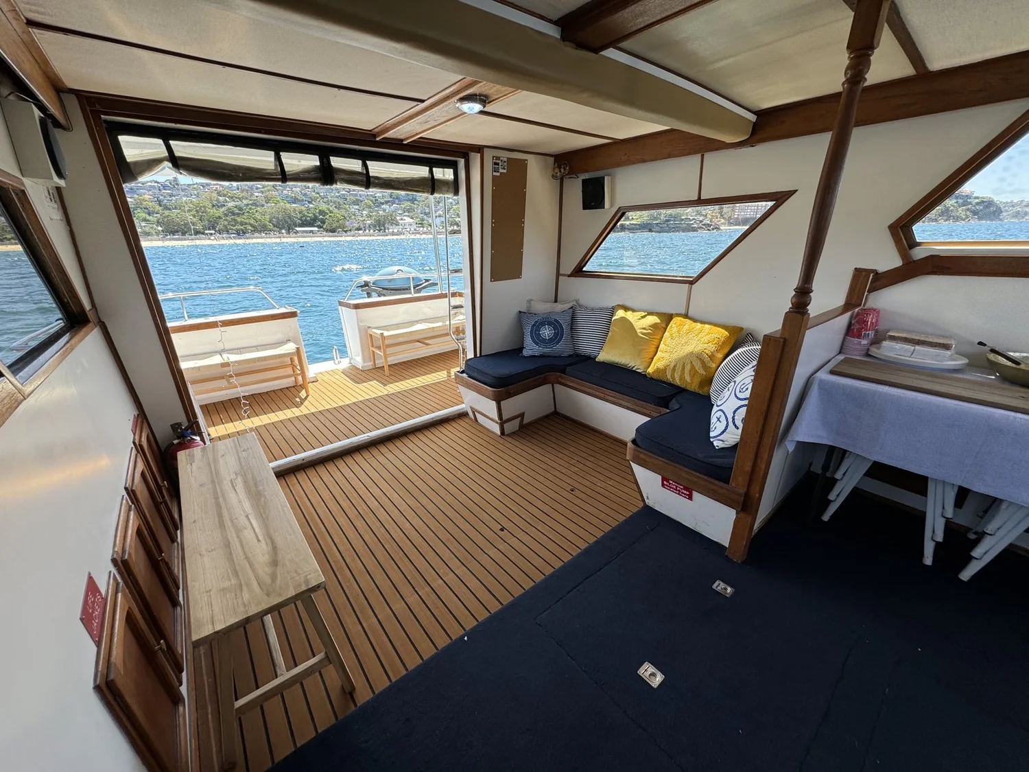 The saloon on Mayfair, a cushioned lounge and a dining table looking out over the timber aft deck to Sydney Harbour