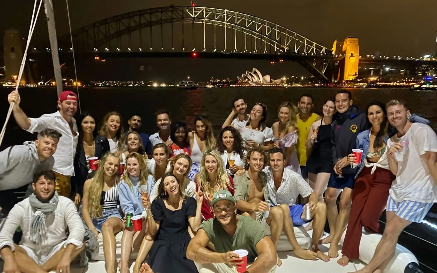 Guests celebrating on the deck of Barefoot Spirit at night on Sydney Harbour, with the Harbour Bridge and the Opera House lit up behind