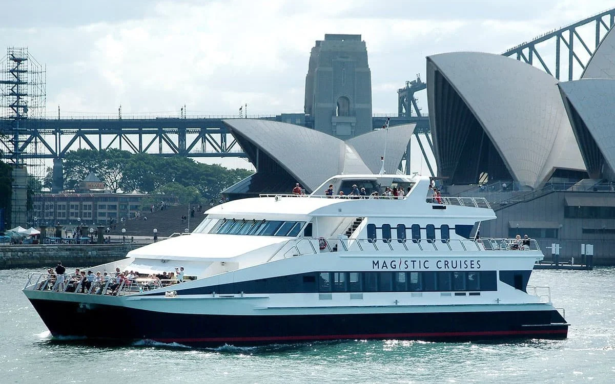 Magistic Two, a white three-deck luxury catamaran, cruising past the Sydney Opera House and Harbour Bridge