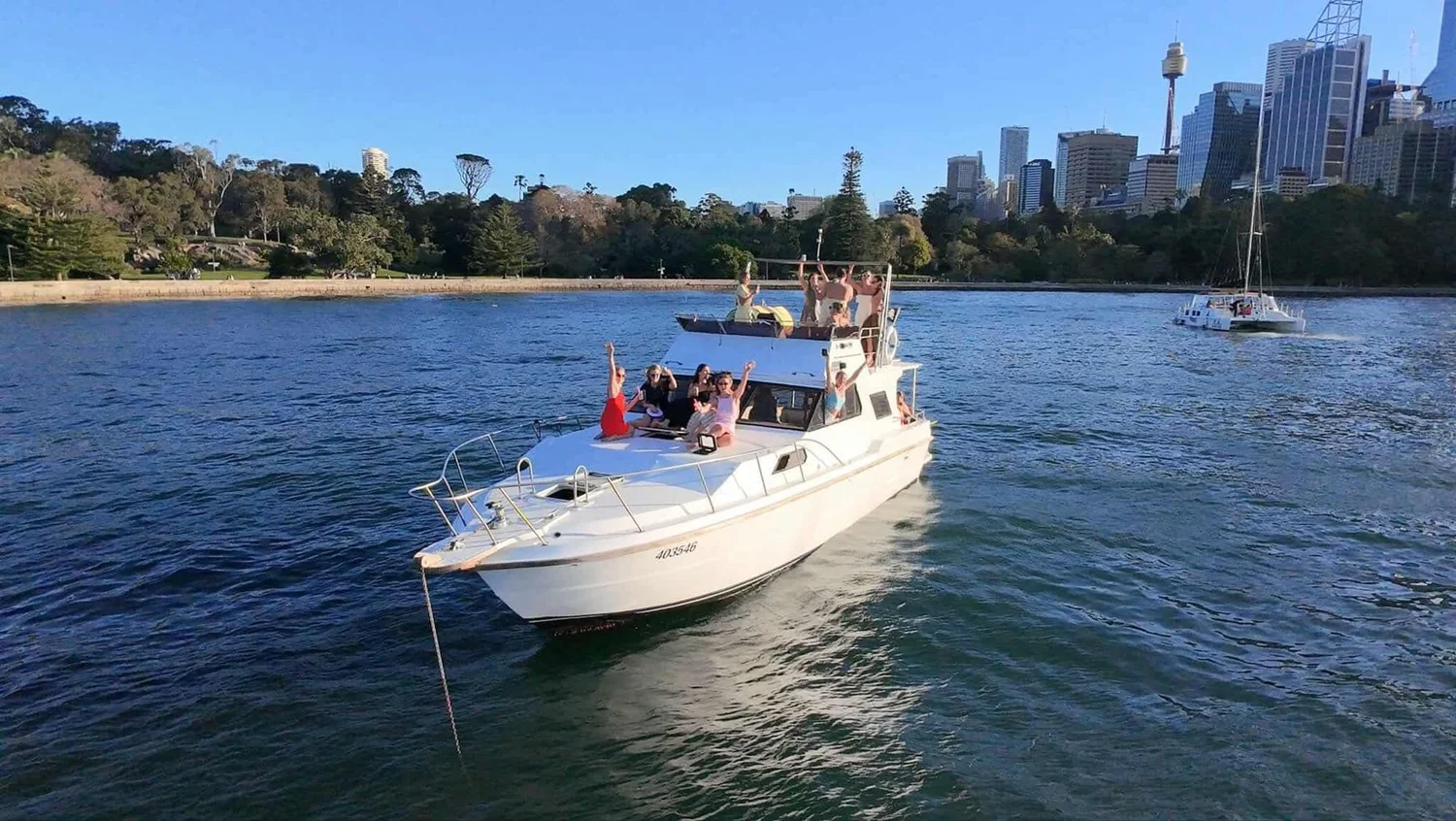 Mayfair, a 43ft motor yacht, at anchor on Sydney Harbour with charter guests waving from the decks and the city skyline behind
