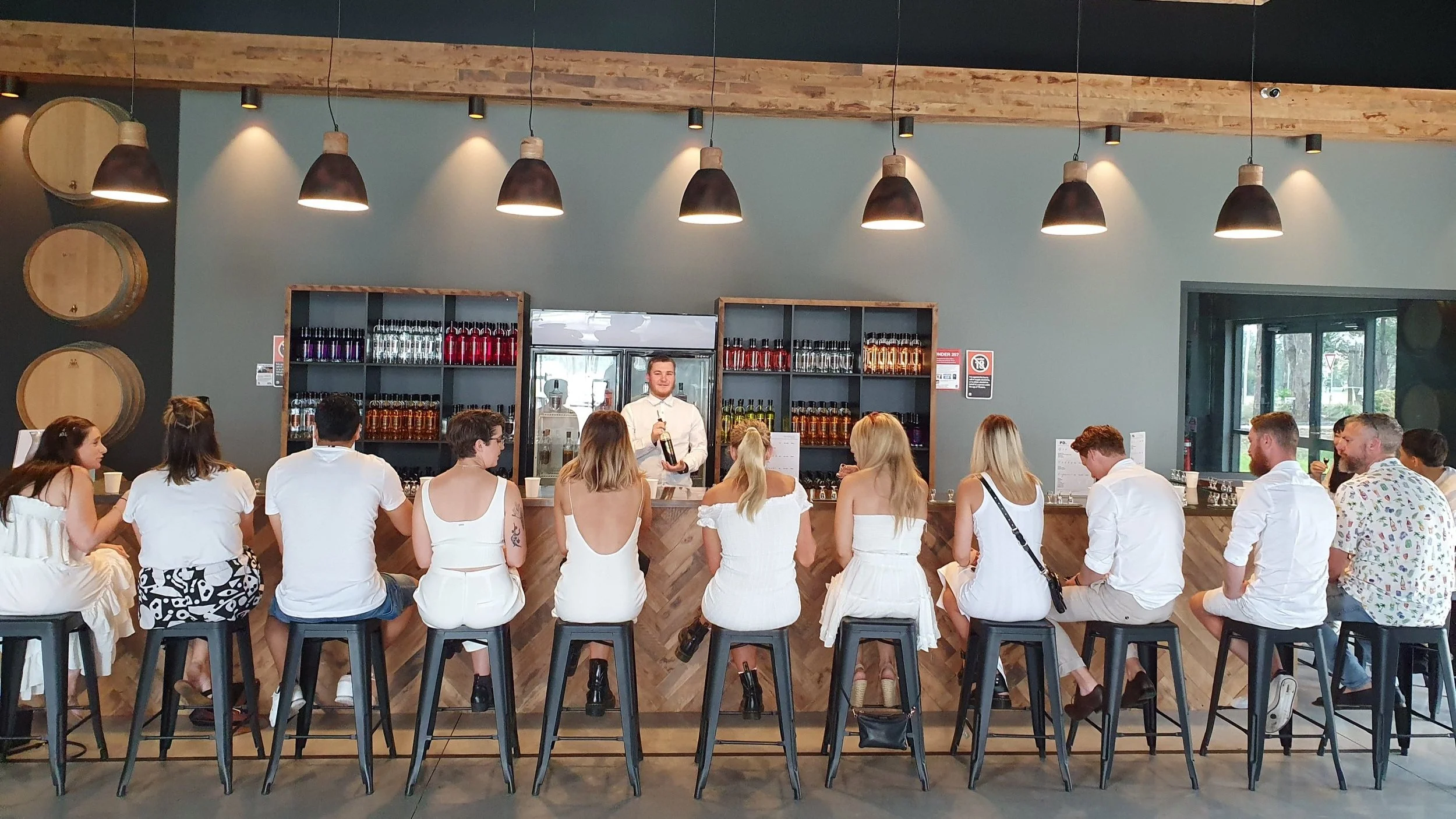 Around fifteen guests seated in a single row along the tasting bar at a Hunter Valley distillery, barrels on the wall behind and a host pouring, on a private brewery and distillery tour.