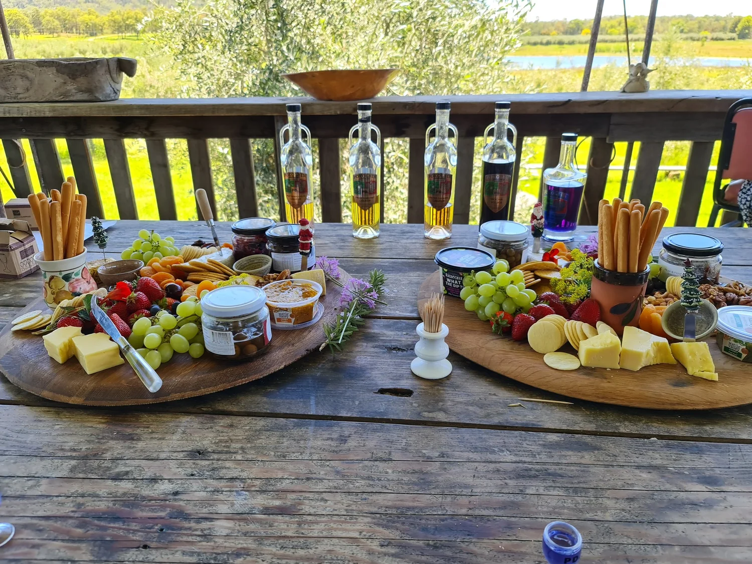 Two large round grazing boards loaded with cheeses, crackers, grapes, strawberries and dips on a rustic timber deck, olive grove behind, on a Hunter Valley wine tour.