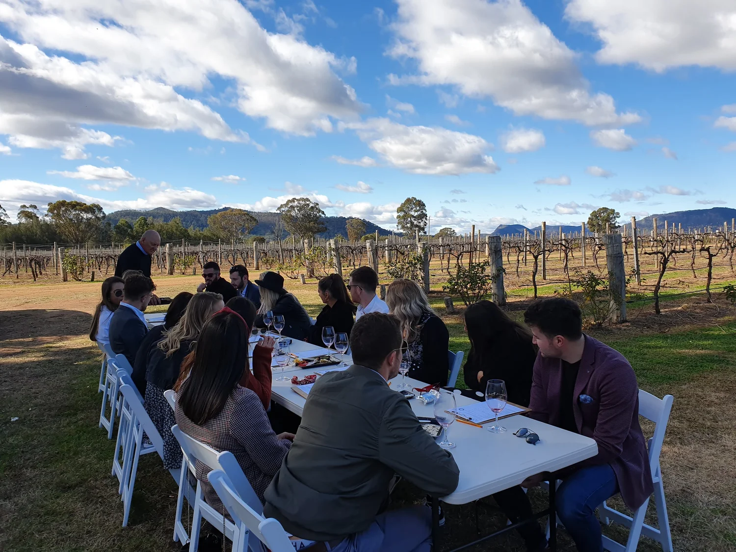 Around eighteen corporate guests seated at long tables set out among the vine rows for a boutique wine tasting, Brokenback Range behind, on a Hunter Valley wine tour.