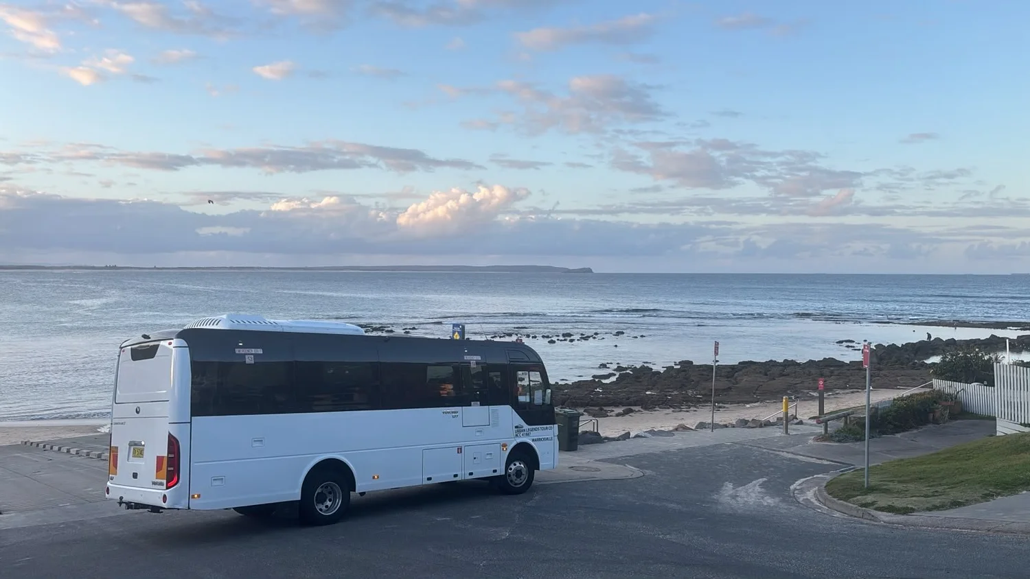 Urban Legends 27-seater coach parked beside the beach at dusk with the rock shelf and headland behind
