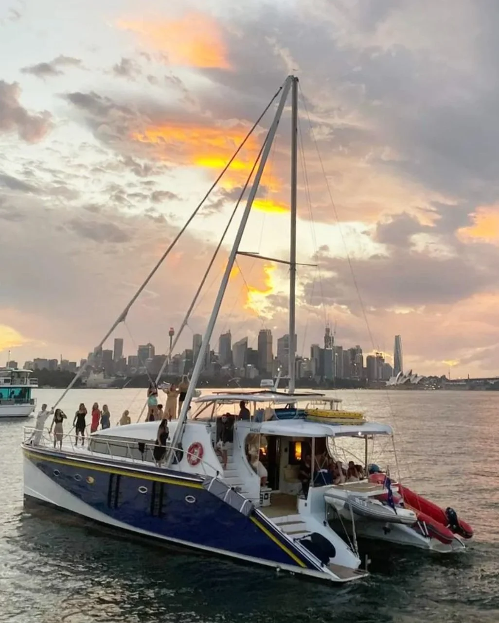 Passion, a dark blue hulled charter catamaran with a red tender alongside, under way on Sydney Harbour at sunset with guests along her rail and the city skyline behind
