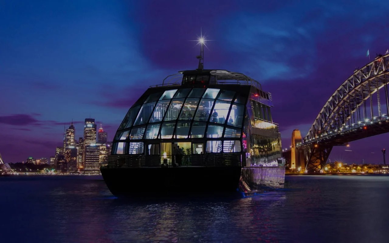 Clearview, a glass-walled event boat, lit up at dusk on Sydney Harbour beside the Harbour Bridge