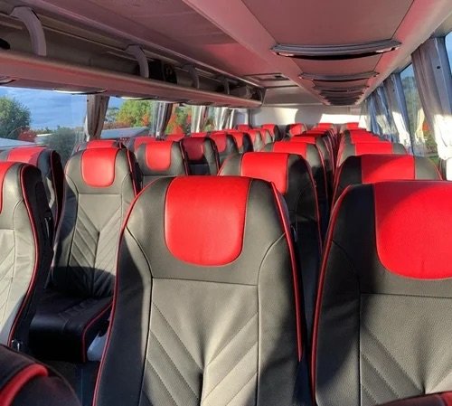 Inside a 57-seat full-size touring coach looking down the aisle, cream and red leather reclining seats with overhead lockers either side