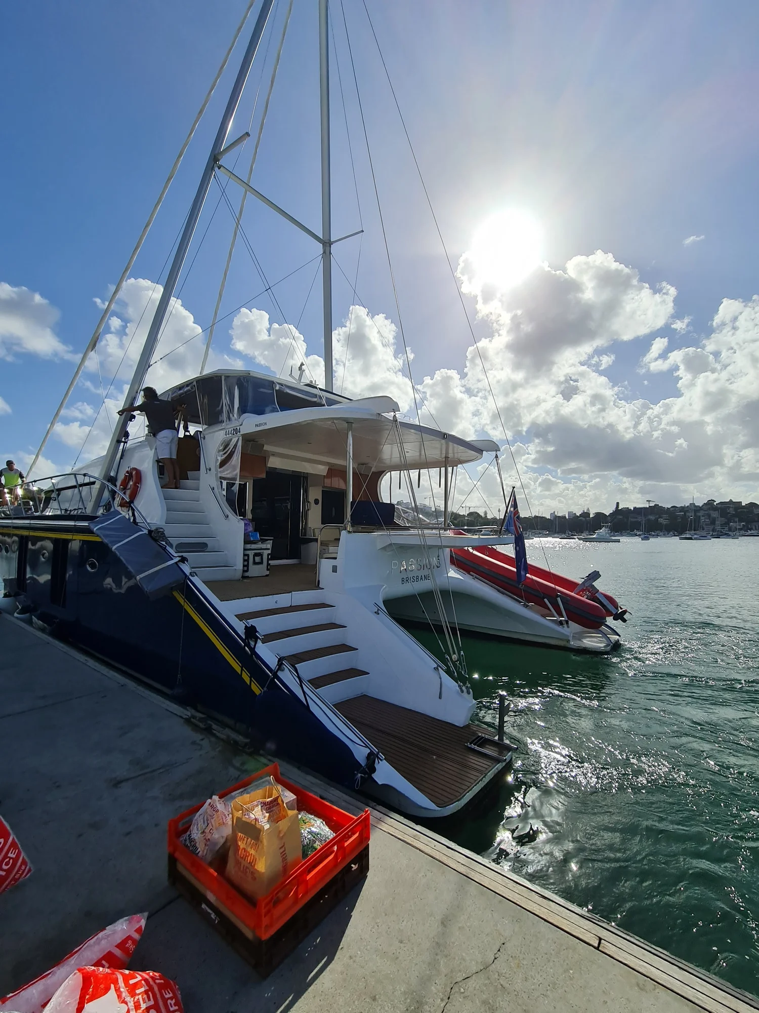 Crates of food and drinks stacked on the wharf beside the boarding steps of a moored charter catamaran, waiting to be loaded before guests arrive