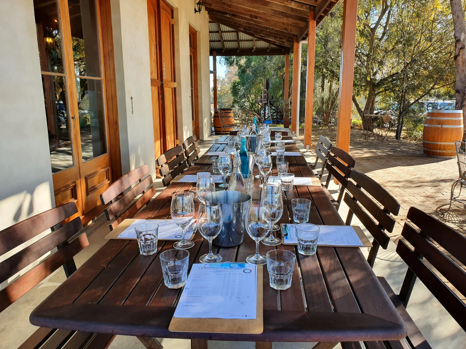 A long timber table set with tasting mats, glasses and water on a shaded cellar door verandah, ready for a seated Hunter Valley wine tasting.
