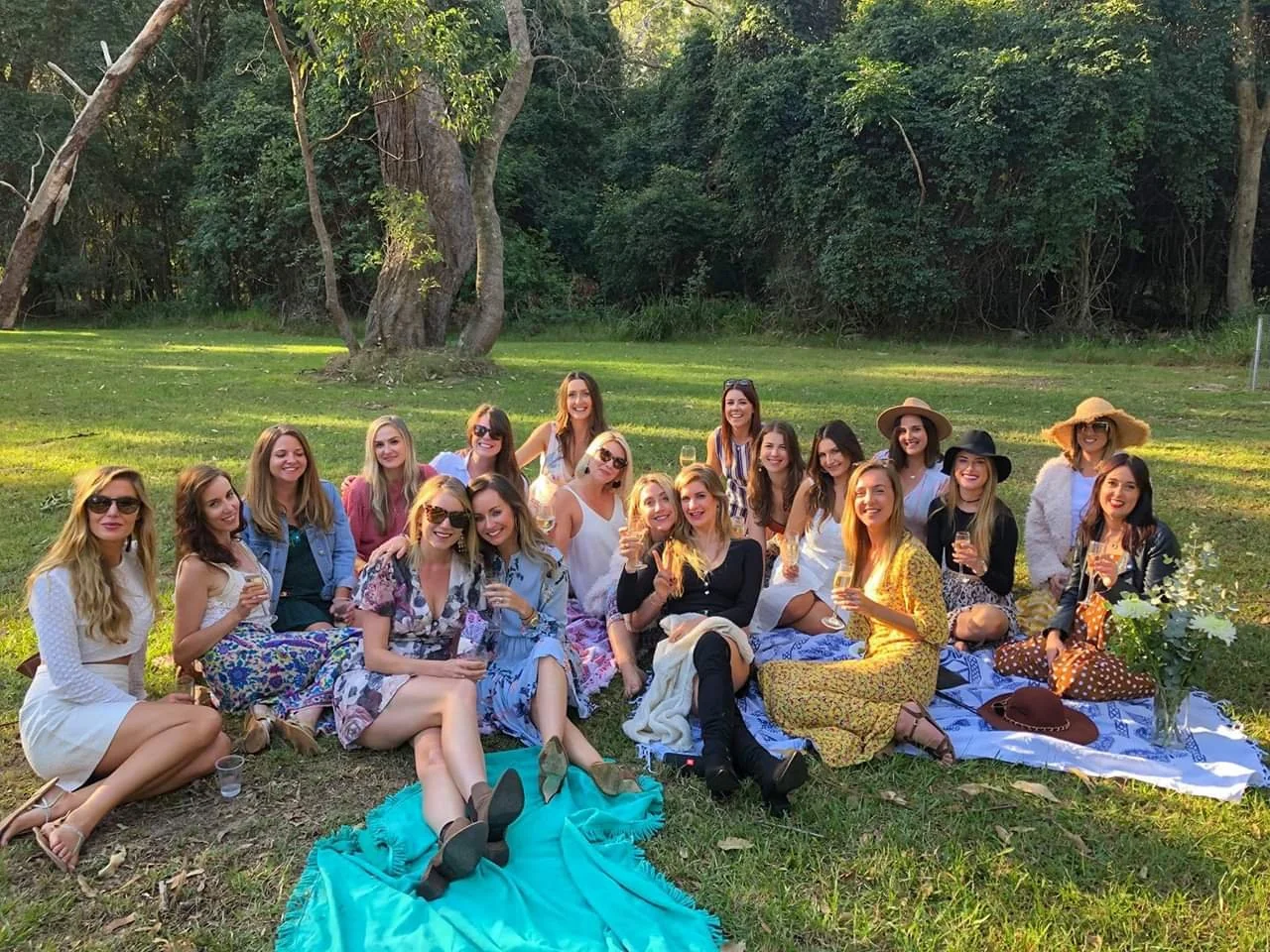 Around twenty two guests seated together on picnic rugs with sparkling wine under a gum tree at a Hunter Valley winery on a hens party wine tour.