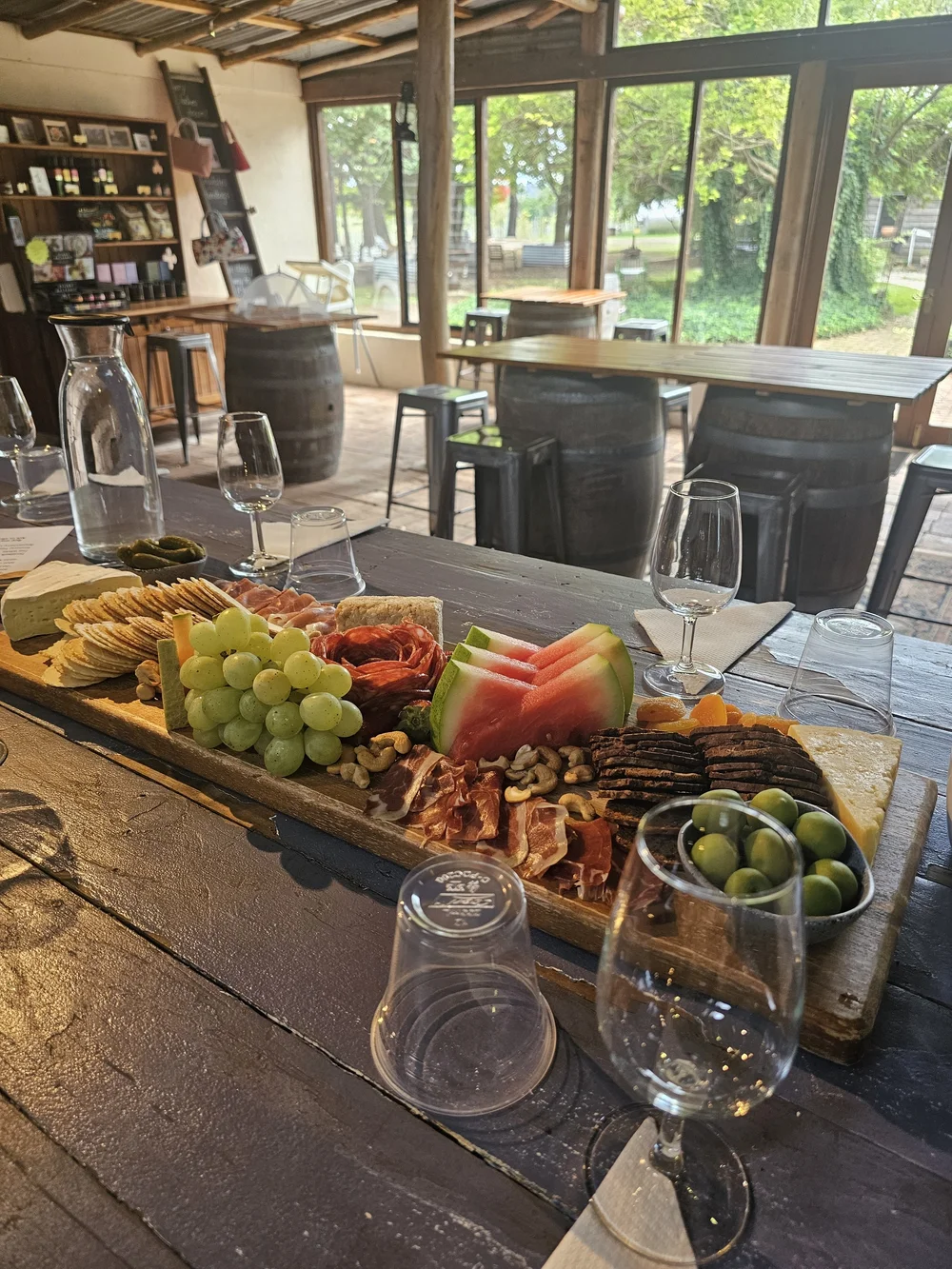 Seated tasting and grazing board on the Southern Highlands 3 x Epic Winery Tour A long grazing board of cheese, crackers, grapes, watermelon and cured meats on a cellar door table, tasting glasses and wine barrels behind it.