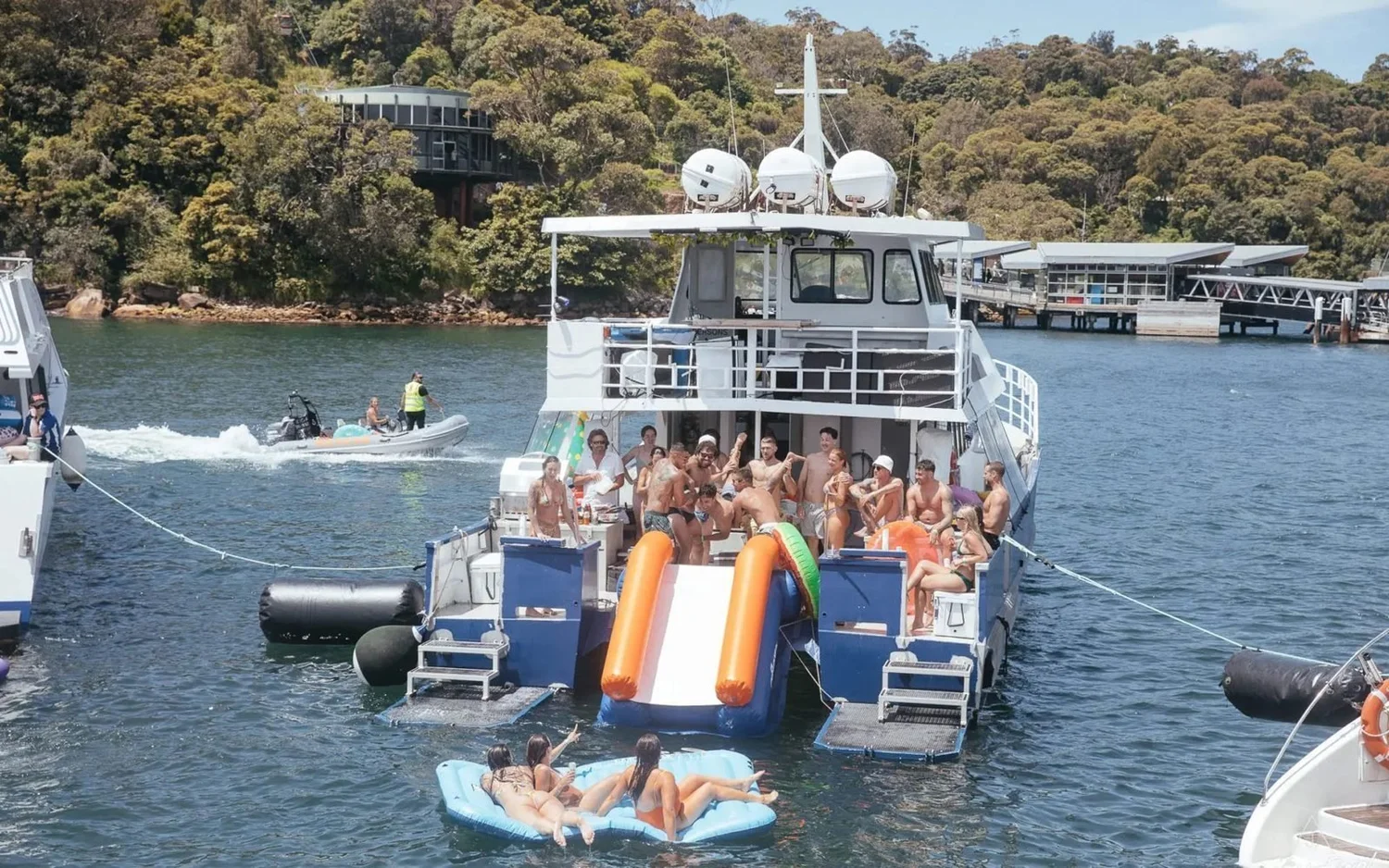 Barefoot Blue at anchor in a Sydney Harbour bay with guests on the stern, the waterslide down to the water and floats alongside