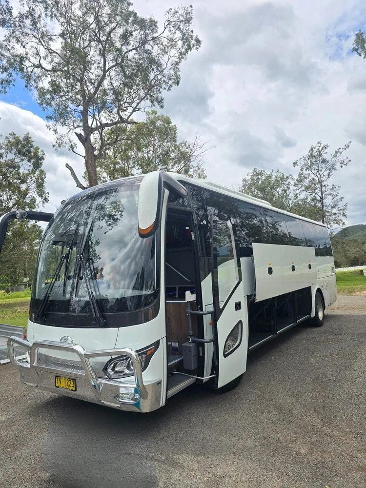 Urban Legends 41-seat luxury coach, plain white with a silver metallic bull bar, parked on grass with gum trees behind before a Sutherland Shire group charter