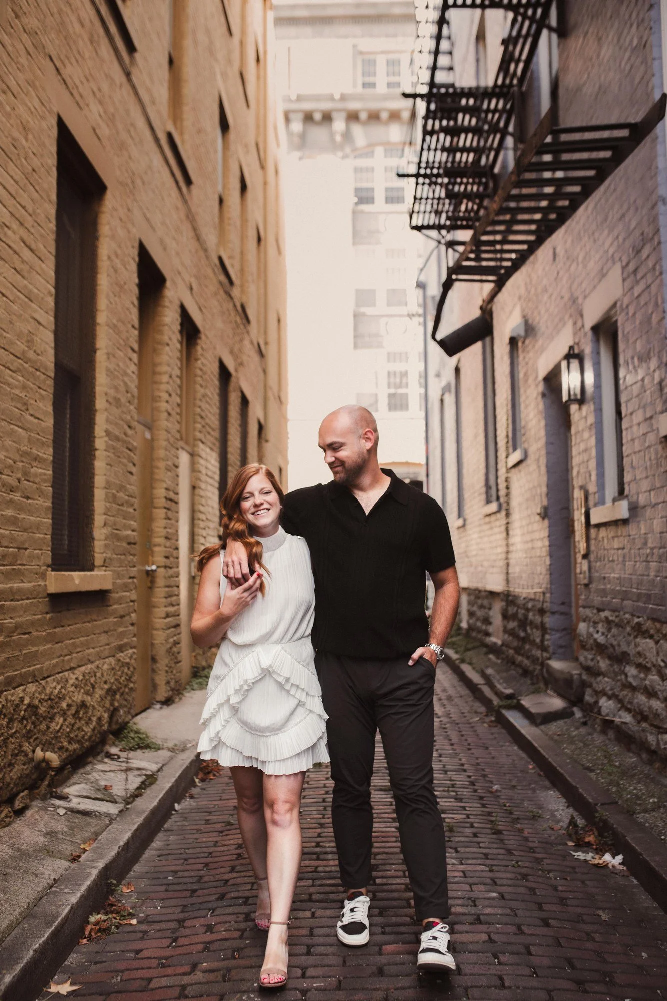 Jacob and Erin holding hands in a brick alley beneath fire escapes.