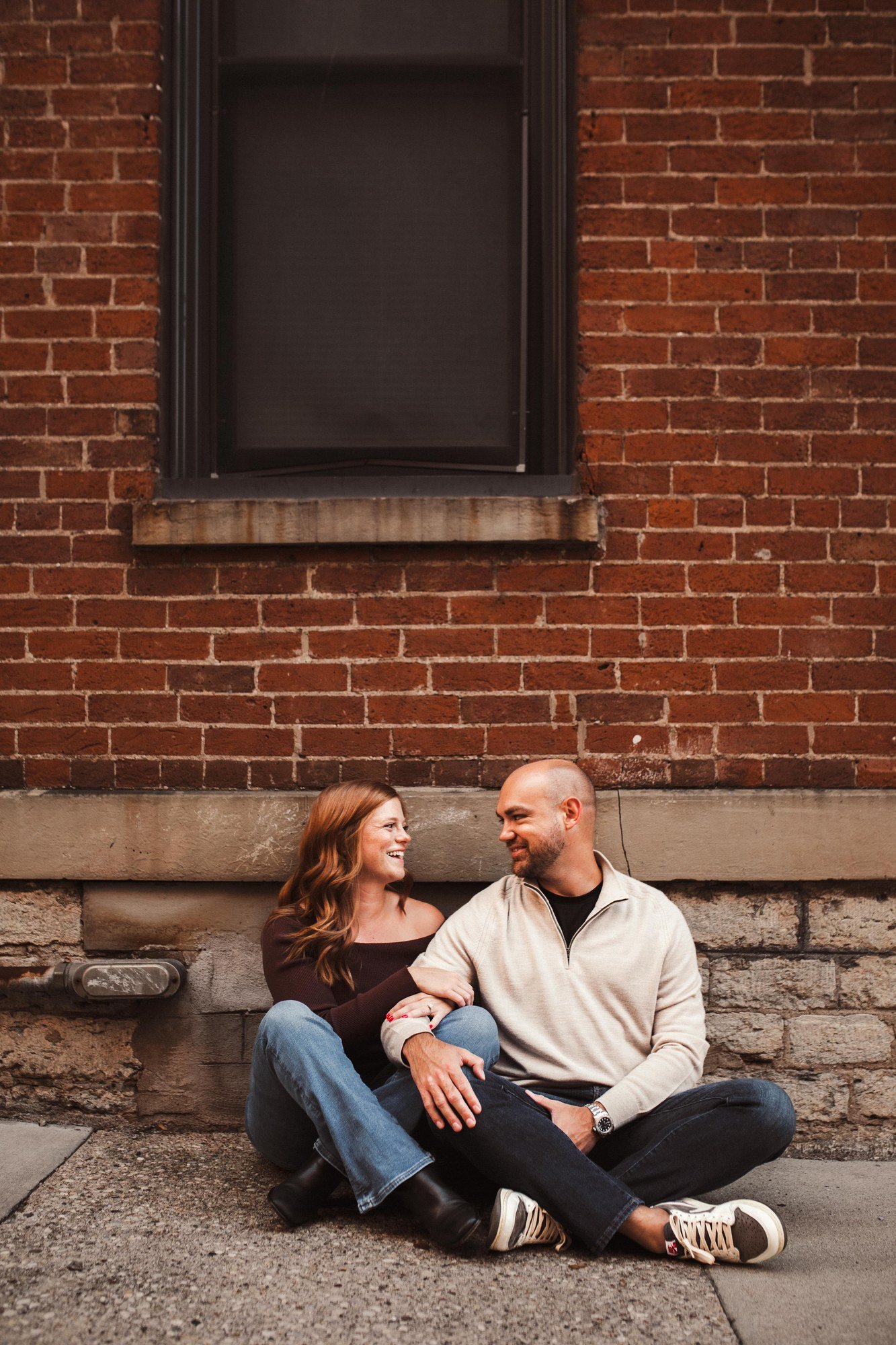 Jacob and Erin seated together beneath a window in a red brick wall.