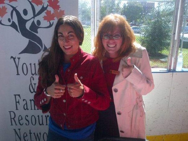 Allison, a glasses-wearing redhead & friend Marianne, a brunet with long hair, smiling with thumbs up at a United Way Windsor-Essex event.