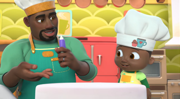 A picture of Cody, from Cocomelon Lane, and his dad. Cody is a young black boy and his dad is a black man with a goatee. They are both wearing aprons and chef's hats. The dad is holding icing to draw on a white cake.