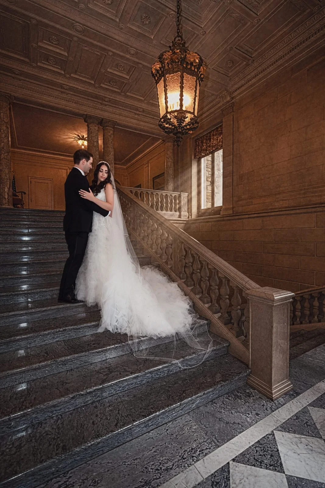 Aldrich Mansion wedding videographer, Warwick, RI — wide portrait of the bride and groom on the marble staircase beneath the gilded lantern chandelier