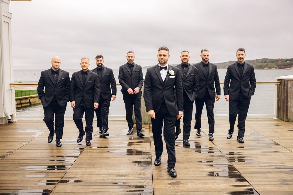 Beauport Hotel wedding photographer, Gloucester, MA — groom and groomsmen walking the rain-soaked harborfront deck with Gloucester Harbor behind them