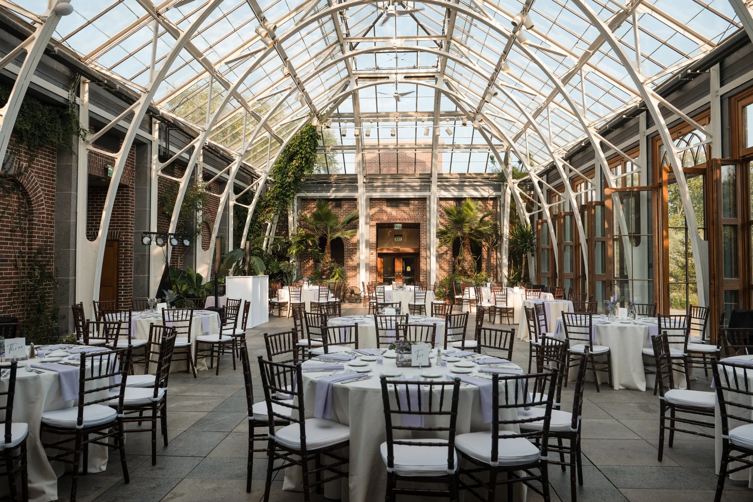 Wide interior view of the Orangerie glass conservatory set for a wedding reception with round white-linen tables, floral centerpieces, and wooden cross-back chairs at Tower Hill Botanic Garden, Boylston, MA