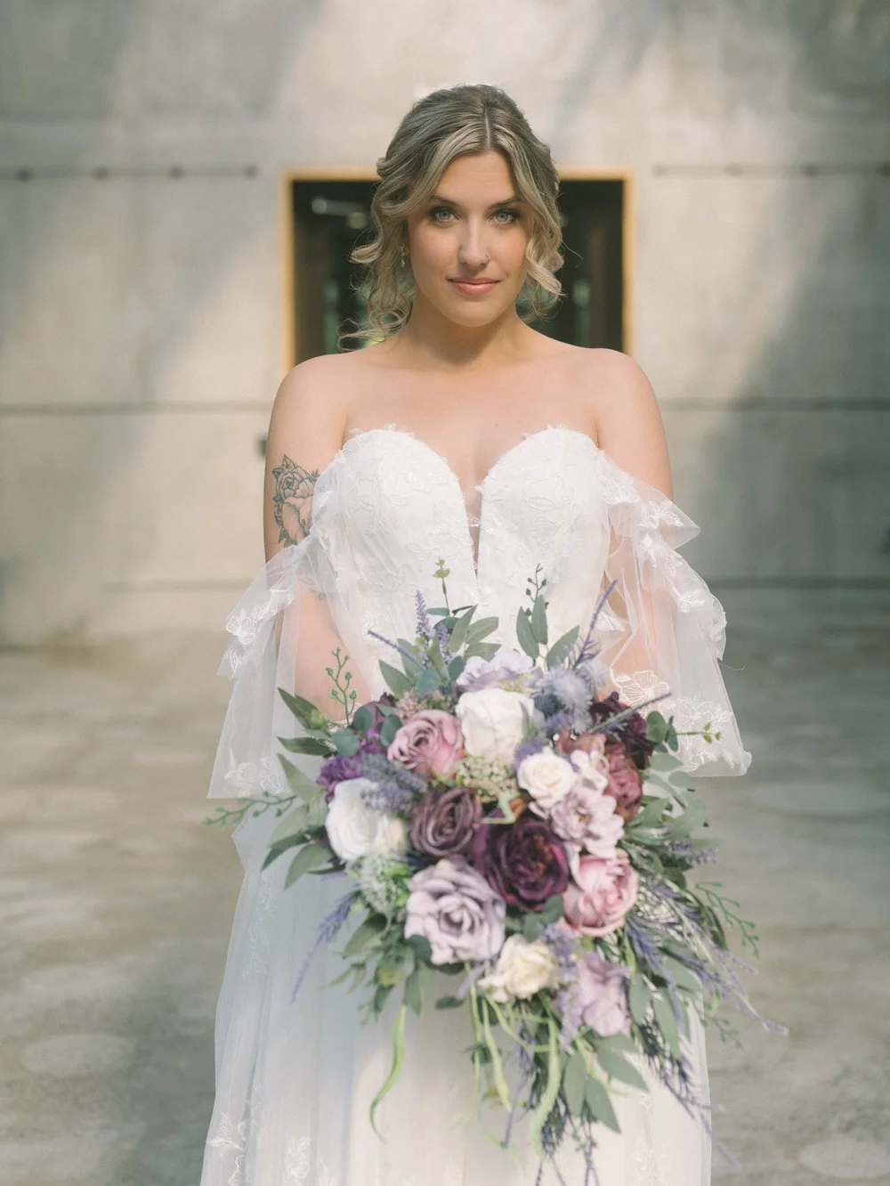 Bridal portrait holding a purple and lavender bouquet against the conservatory stone wall at New England Botanic Garden at Tower Hill, Boylston, MA