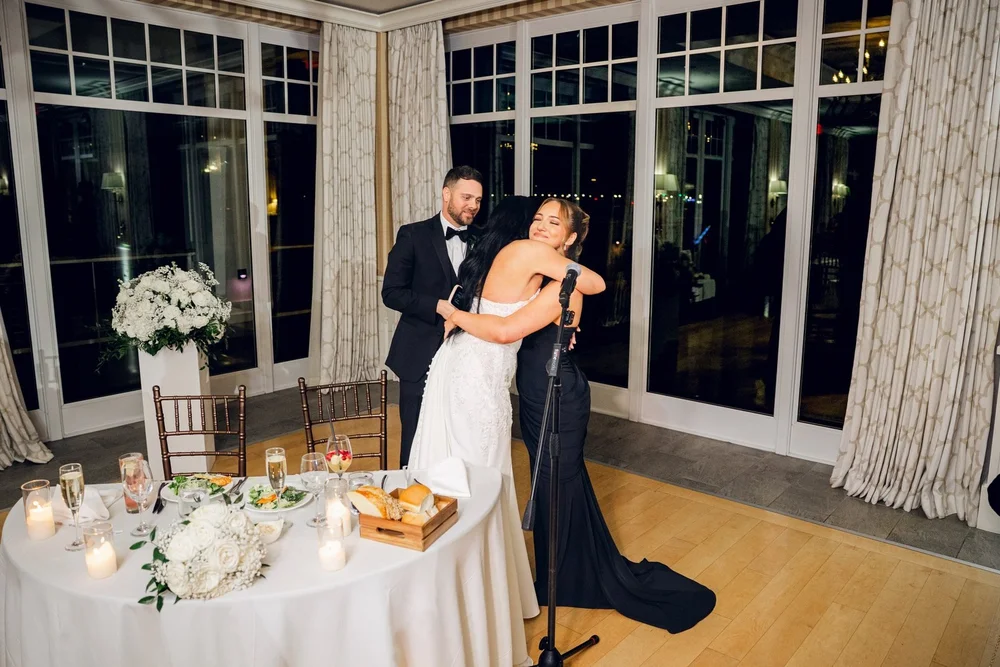 Beauport Hotel wedding videographer, Gloucester, MA — bride embracing a toast-giver at the sweetheart table framed by harbor-view windows