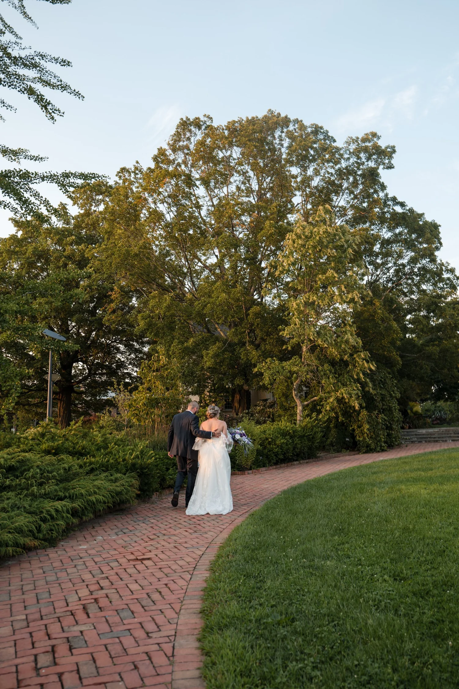 Wide vertical portrait of the bride and groom walking along a curved brick pathway beneath towering shade trees and manicured lawns at Tower Hill Botanic Garden, Boylston, MA